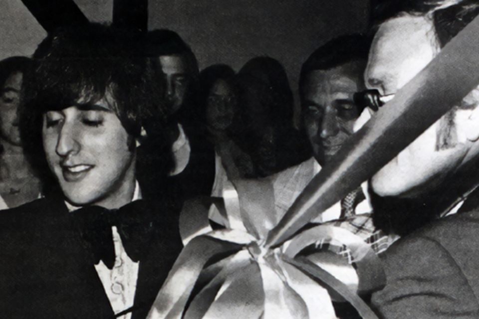 Black and white photo of a man in a tuxedo cutting a ribbon as others look on. 
