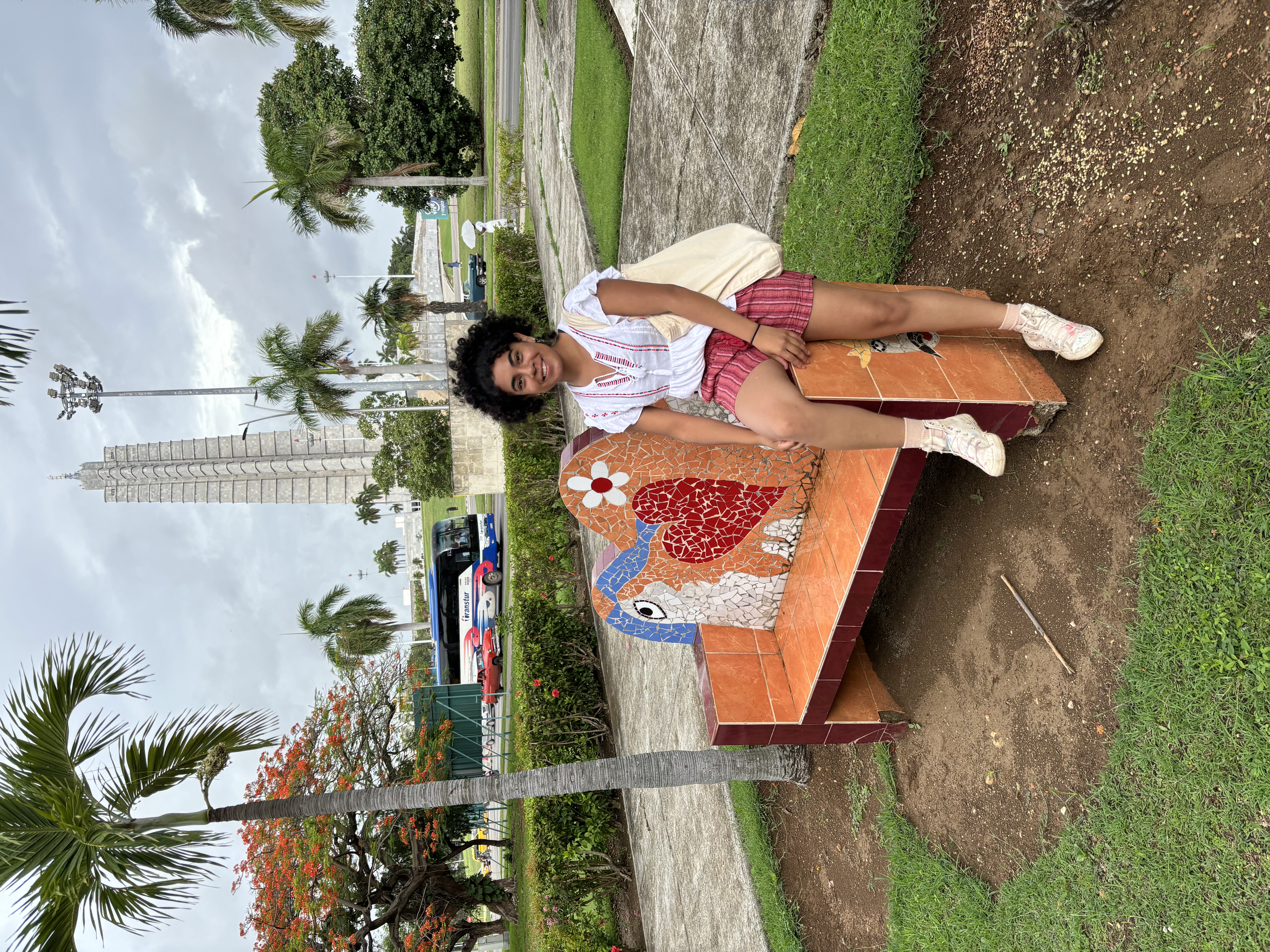 Angelica perched on a bench in a park in Mexico, with buildings and palm trees in the background. 