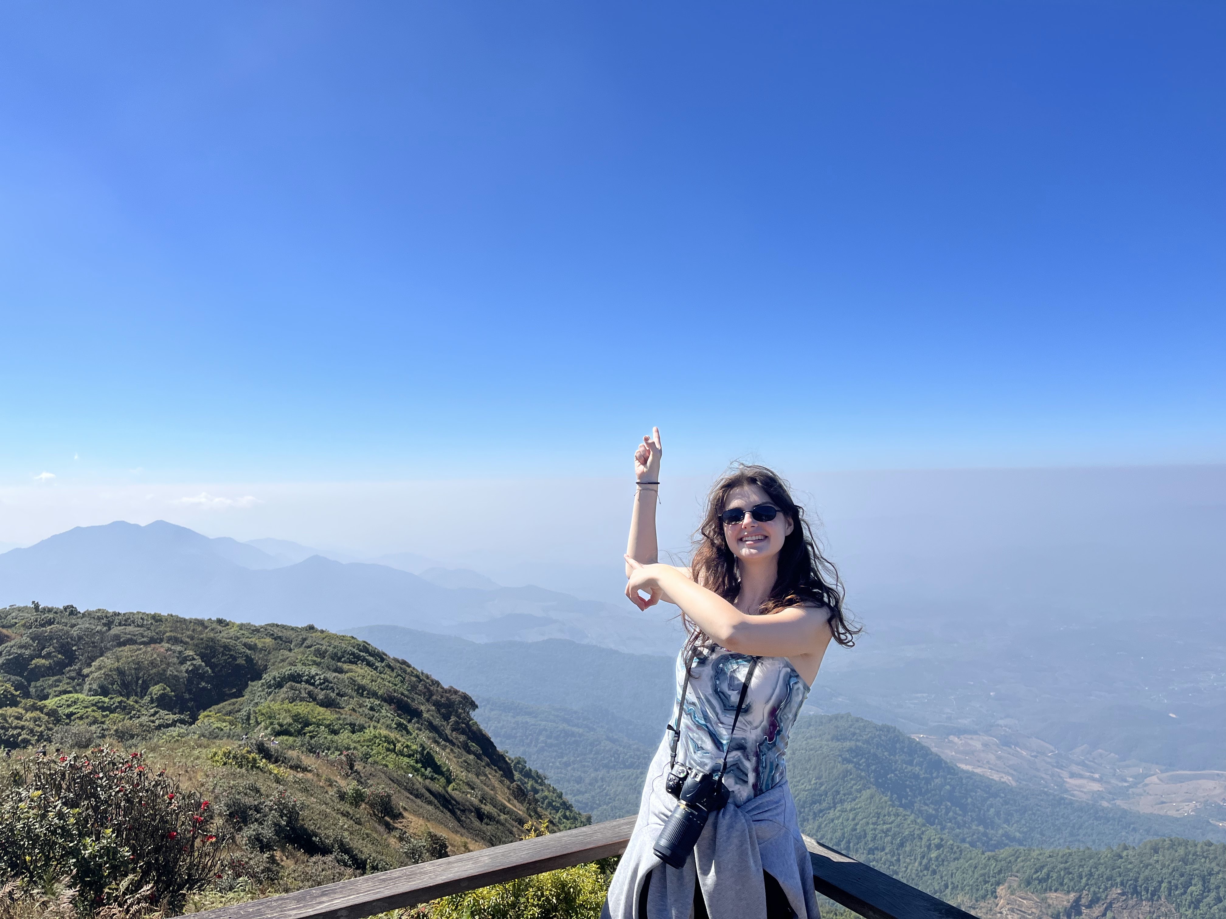 Hannah stands at a precipice, pointing in the air at a mountain in the distance, with blue sky all around her.