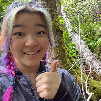 A headshot of Natalie. A female with pink and purple hair braided. She is wearing a dark colored jacket and she is giving a thumbs up.