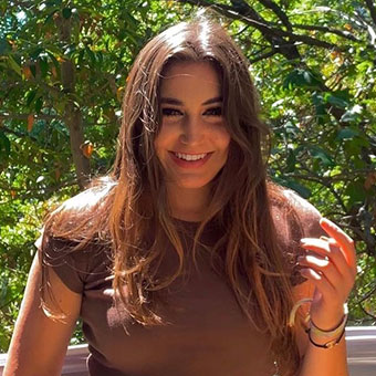 A headshot of Alex. A female with long brown hair wearing a brown shirt and a blue printed skirt. She is standing outside.