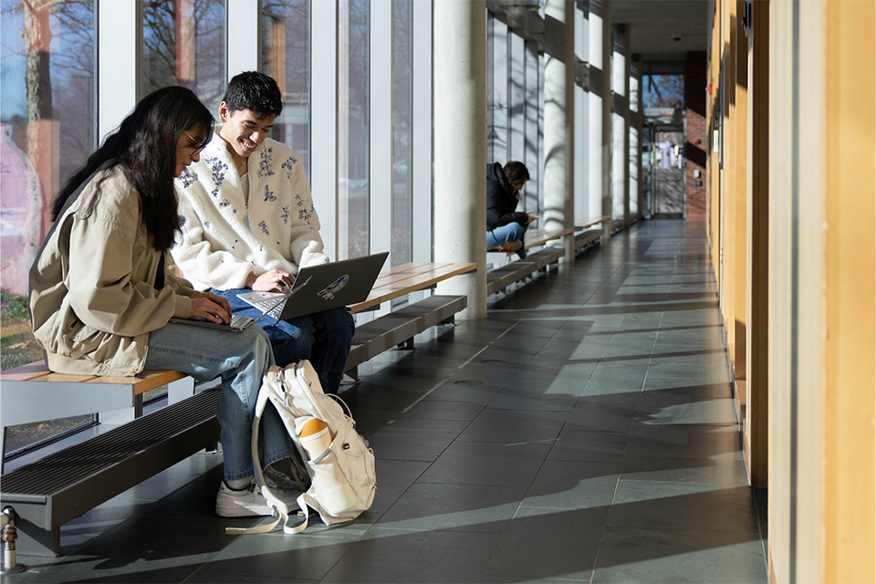 Two students sitting on a bench in a hallway in front of a floor to ceiling window