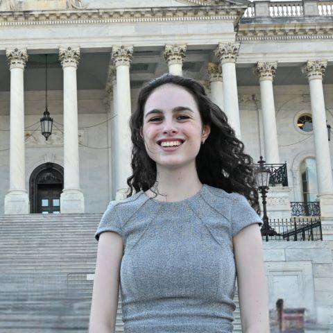 Mandy stands in front of a white marble building. She has long brown hair and wears a grey short sleeve sweater.