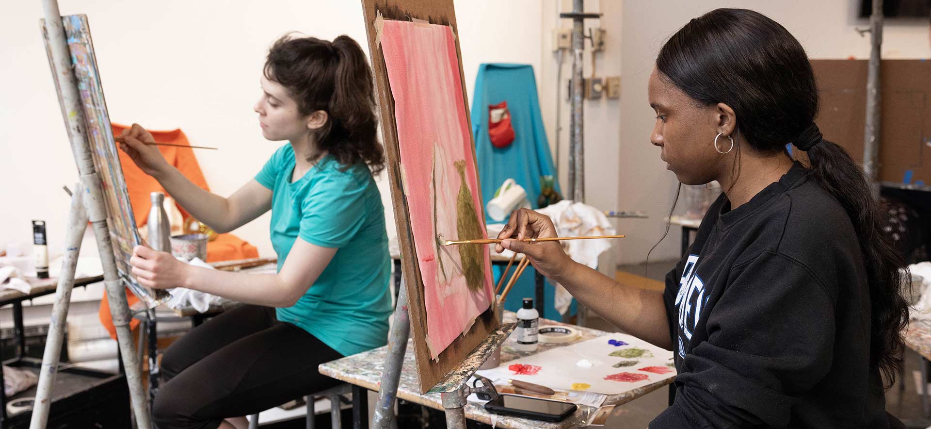 Two people sit on stools and paint on canvases