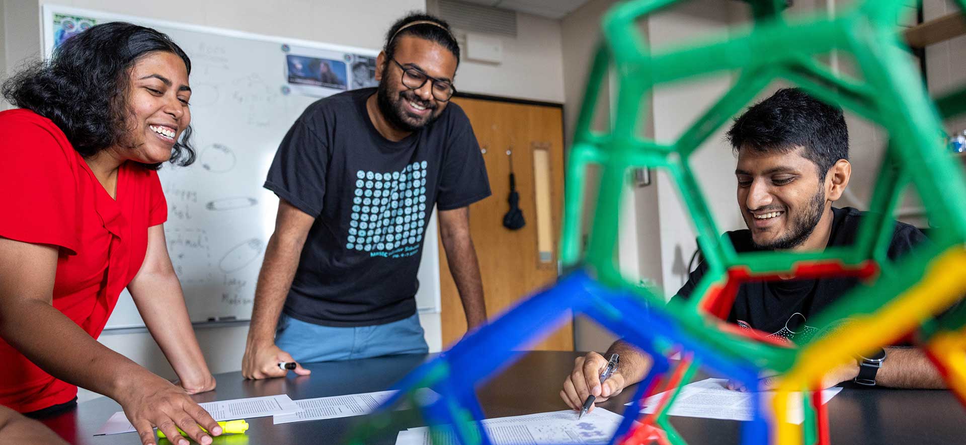 Graduate students work together in a classroom. One is viewed through a 3D geometric structure
