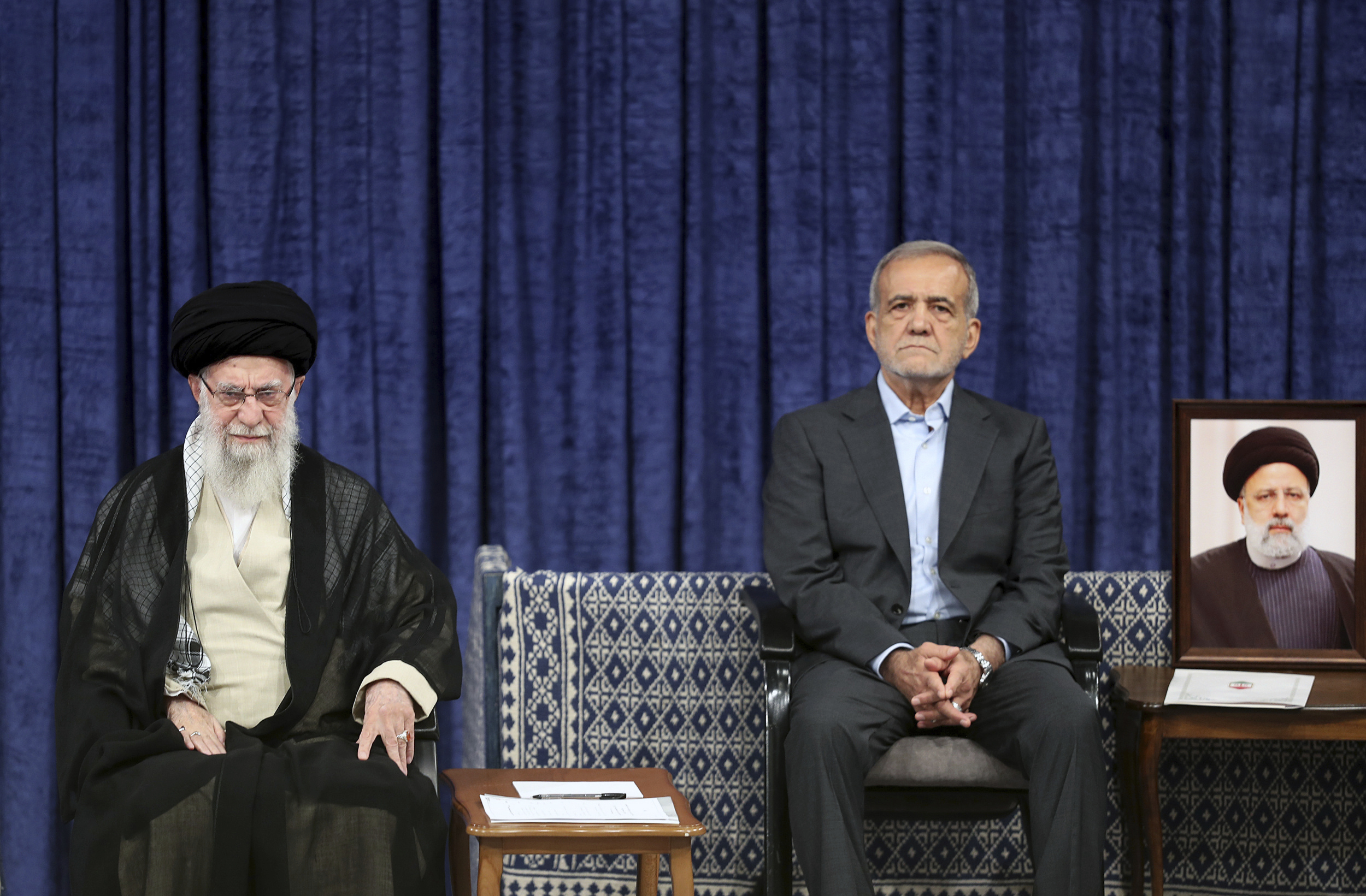 Iran's Supreme Leader Ali Khamenei and President Masoud Pezeshkian sit on a stage at a ceremony in Tehran, Iran. Next to Pezeshkian is a framed portrait of a cleric propped on a small table.