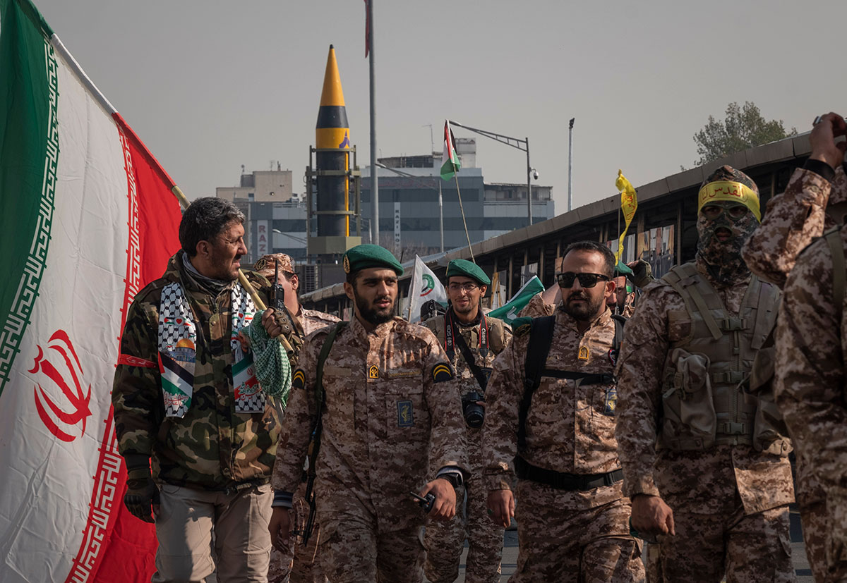 A group of military personnel walk at a military rally in Tehran, Iran, in November 2023. One holds a large Iranian flag while missiles are unveiled in the background.