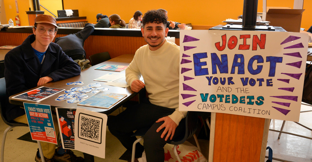Two students at a cafeteria table with an ENACT Your Vote Day of Action sign