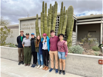 Brandeis students and faculty stand with a Park Ranger in front of a large cactus at the entrance to the Organ Pipe Cactus National Monument sign and building.