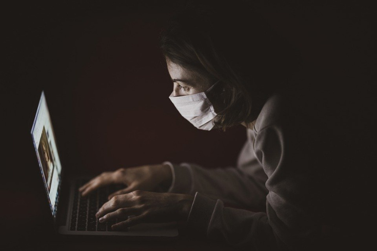 Person wearing a mask, working on a laptop in the dark.