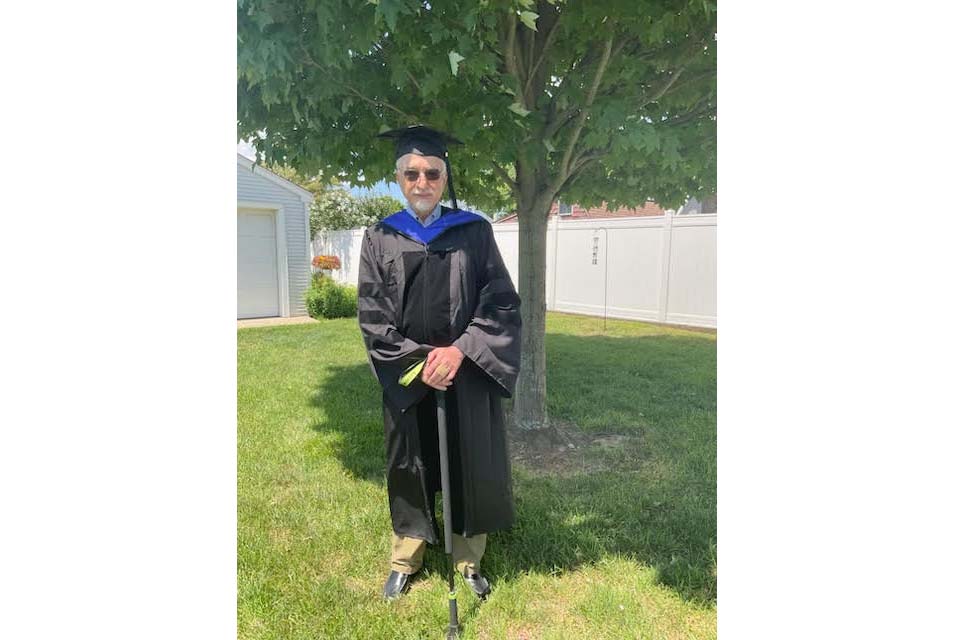 Bill Abbate, wearing graduation robes and holding a cane, stands in front of a tree.