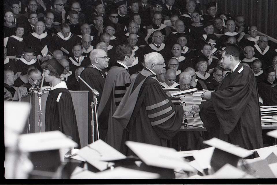 A university official shakes hands with and hands a diploma to a graduating student in cap and gown as robed faculty members look on.