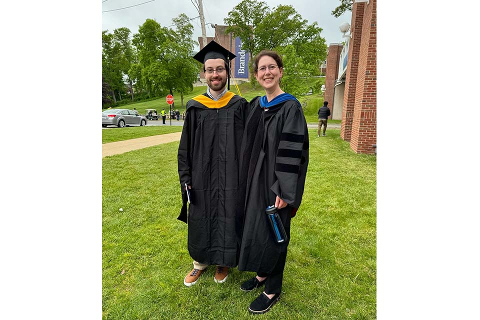 Josh Broderick Phillips and Lotus Goldberg stand on the Brandeis campus wearing academic regalia.