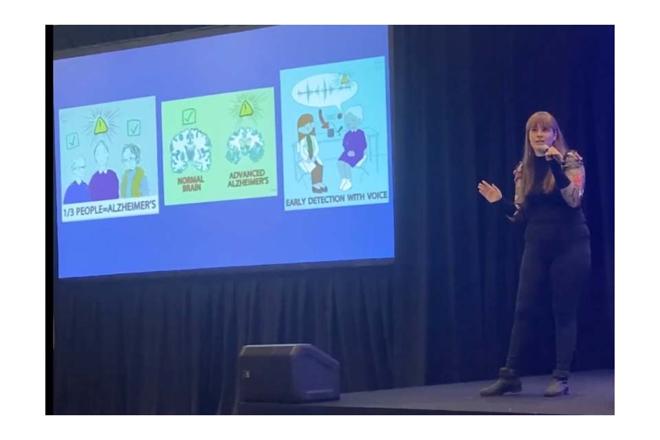 Liz Mahon stands onstage, speaking into a microphone in front of a projected slide showing cartoon images related to Alzheimer's.