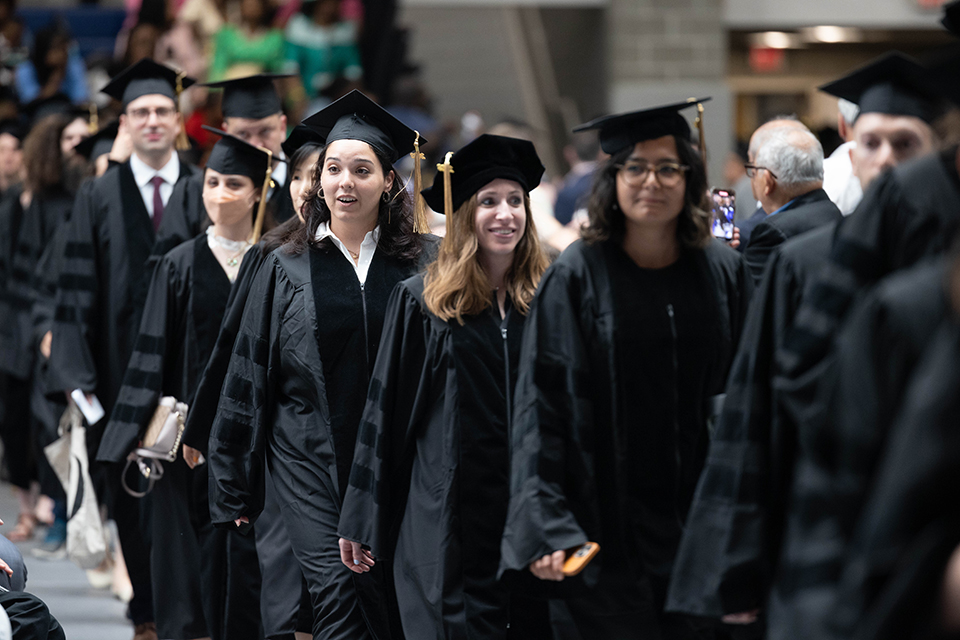 A line of students in black graduation robes and caps walk in the Gosman gymnasium.