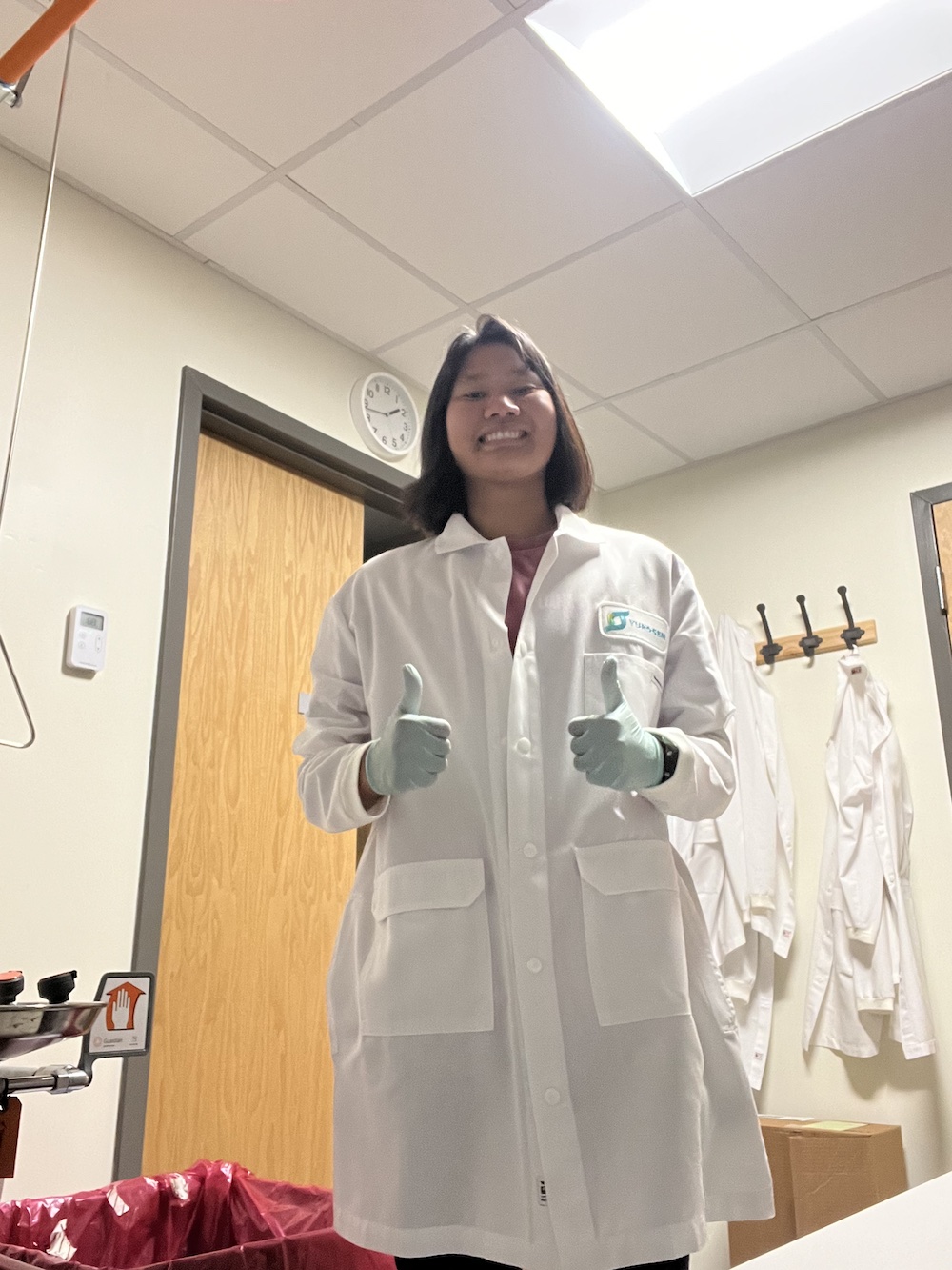 Sydney Lenhart stands in a lab, wearing a lab coat and giving 2 thumbs up.