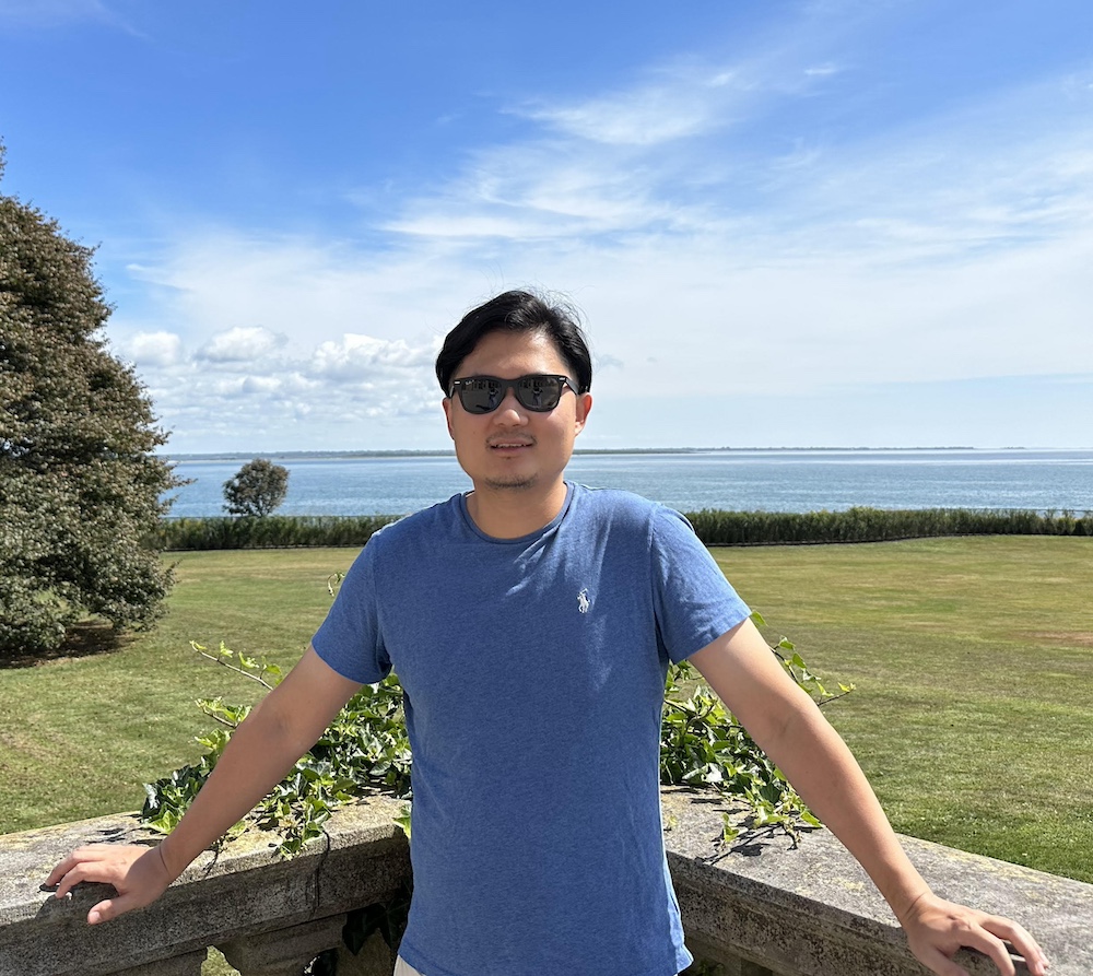 Zhaoxi Zheng stands by a wall in front of a green field and blue sky.