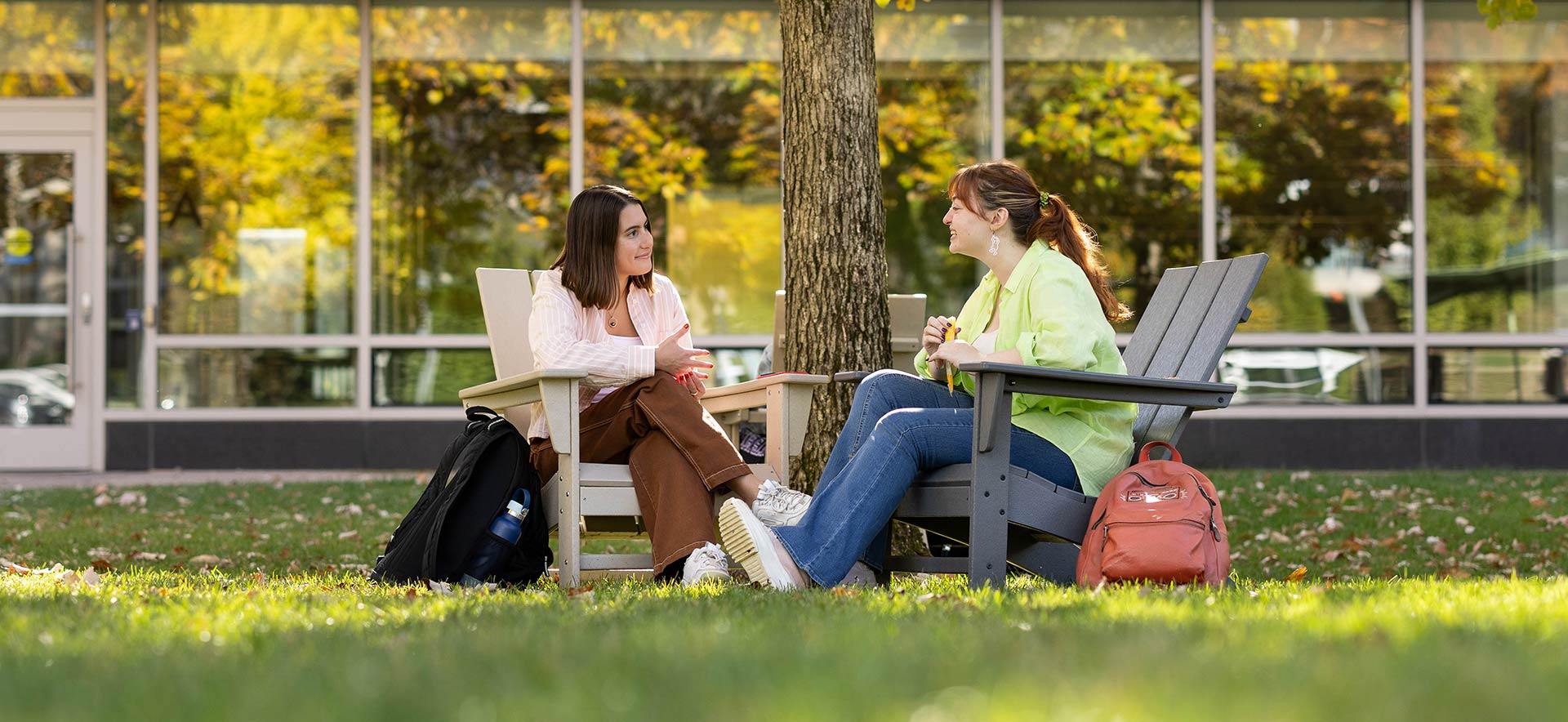 Two people sit outside chatting. A building is seen in the background along with a tree with falling and colorful leaves.