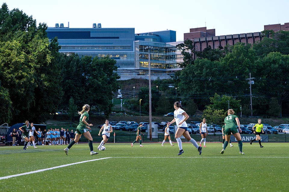 Soccer players on the field with the Brandeis campus in the background