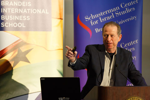 Speaker Erran Carmel at the podium, gestures with his right hand, pointing offstage to his right. 