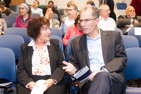 Woman in white and red flowered blouse and dark blazer sits in audience chairs, conversing with tall man who gestures as he speaks.