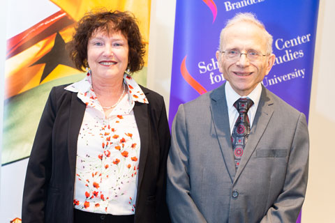 Woman in blouse and blazer and man in grey suit stand smiling in front of colorful banners for the Brandeis International Business School and the Schusterman Center for Israel Studies. 