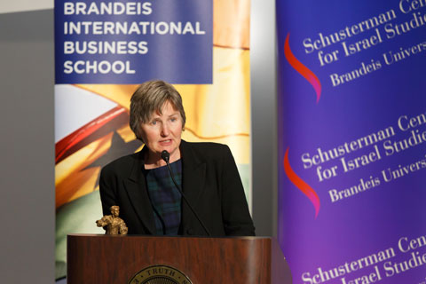 Woman in short grey hair speaks into a microphone at a podium, addressing the off-screen audience, with the colorful banners of the Brandeis International Business School and the Schusterman Center for Israel Studies.