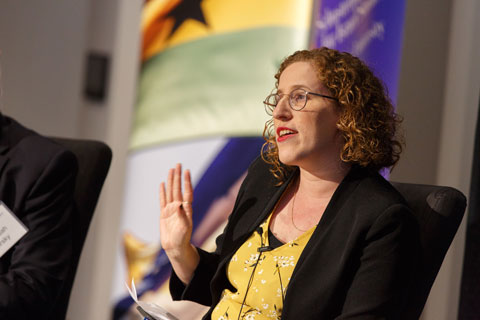 Red-haired woman in yellow blouse and dark blazer raises her hand in a gesture, open palm held vertically, facing the audience, as she speaks. 