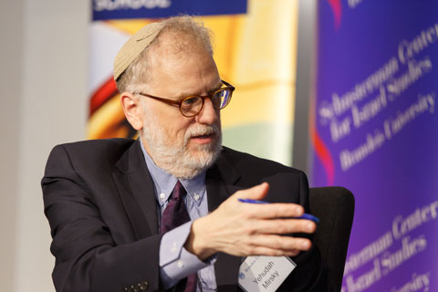 Bearded man in a kippa, also known as a yarmulke, wearing glasses, gestures to listeners outside of photo as he speaks. 