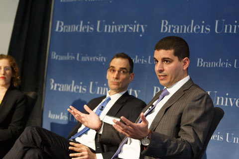 Young man talks to audience (not seen in photo) as an older man in a blue tie seated to the speaker's left listens .