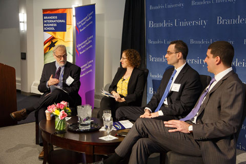 Bearded speaker smiles and gestures toward woman and two women seated near him onstage, as they engage in conversation. 