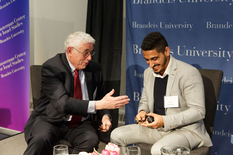 Older, white haired man seated next younger, dark-haired man, gesturing as he speaks, both laughing. 