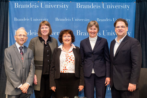 Short, white-haired man, tall, auburn-haired woman, shorter, brunette woman, taller, grey-haired woman, and dark-haired man, stand next to each other, facing the camera, smiling, in front of a blue banner background stamped with the words Brandeis University. 