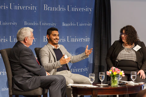 An older man, a younger man, and a young woman seated onstage in discussion, the young man in the middle smiles and gestures as he speaks. 