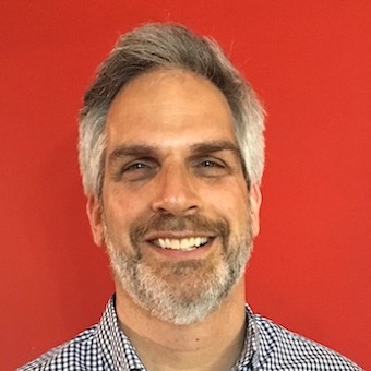 Headshot of Josh Wolk, Journalism Program Professor of the Practice, against a red background wearing a blue and white checked shirt