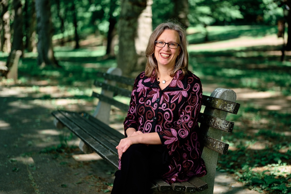 Amy williams, a white woman wearing bright colored clothes, sits outside on a bench