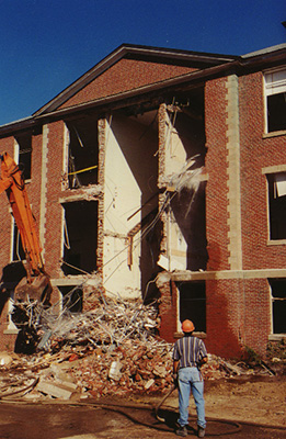 A worker stands in front of the building as a crane pulls debris out from the interior.