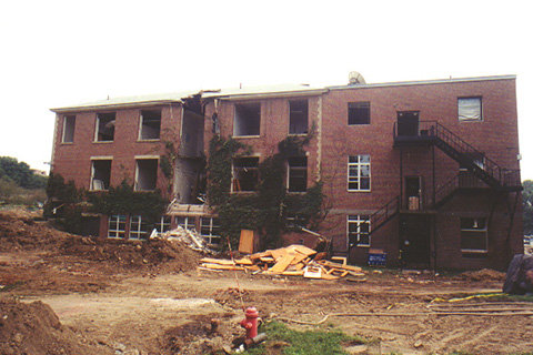 Side of the building with a central section missing.  Debris on the ground and piles of dirt.
