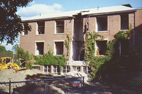View of the side of the building with a central section already torn down.  A bulldozer is towards the back behind a mound of dirt. A chain link fence is in the front.