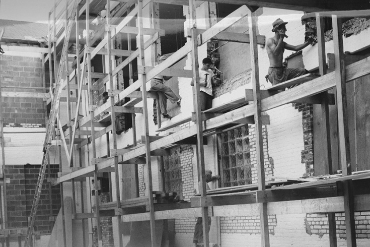 Three workers perched on the scaffolding, using hammers, during the construction of Sydeman Hall