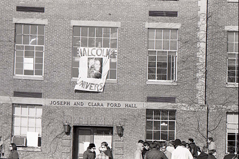 Large fabric sign, hanging outside one of Ford Hall's front windows, stating Malcolm X University with a photo of Malcolm X.