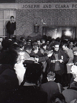 An African American man stands outside of a building at night with a large group of people with microphones and cameras standing around him.