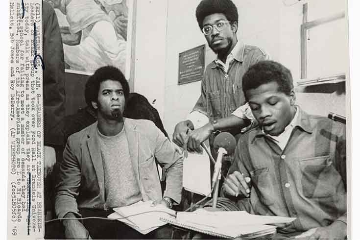 3 young Black men sit in an office space in front a microphone.