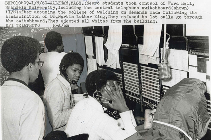 Five African American men inside the communications room in Ford Hall. There is a switchboard and microphone in front of them and a blackboard with writing in white chalk on the wall to the left of them. Three of the men are standing and two of them are sitting and have headsets on their heads. One of the men who is standing is holding a device to his ear.