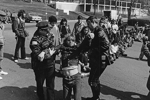 Long group of students sitting and standing on the road and blocking the entrance to campus. Two police officers are holding a man by the arms and dragging him up in order to arrest him. 