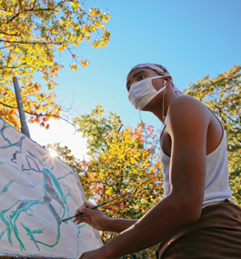 A young man wearing a face mask paints a large watercolor outside in bright sunshine, with the leaves on the trees turning autumnal colors.