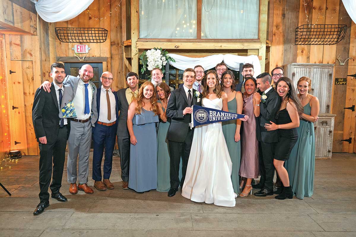 A wedding party holding a Brandeis flag smiles for a group photo.