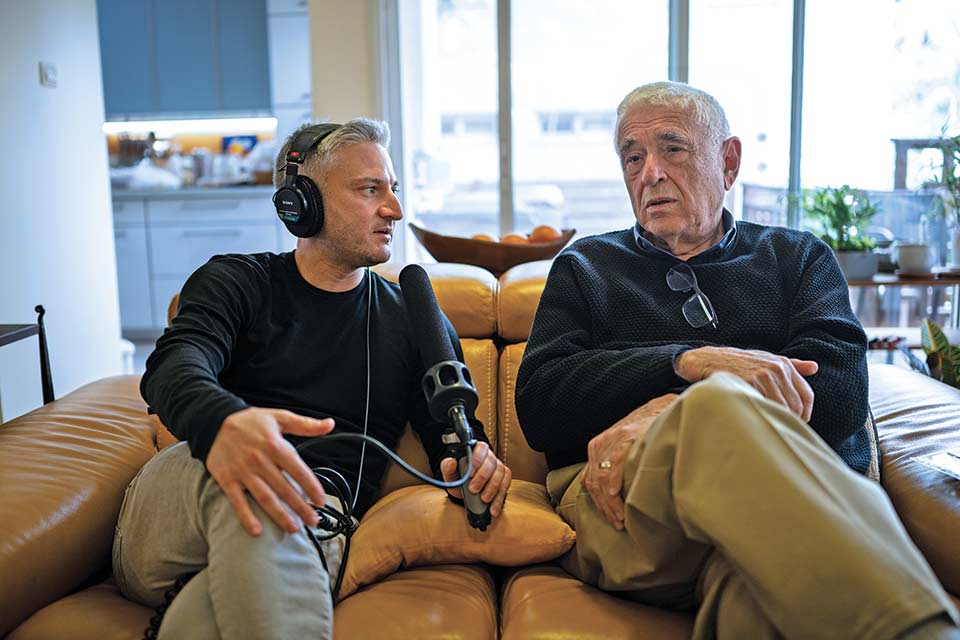 A person in headphones with microphone interviews another person. They are both sitting on a couch.