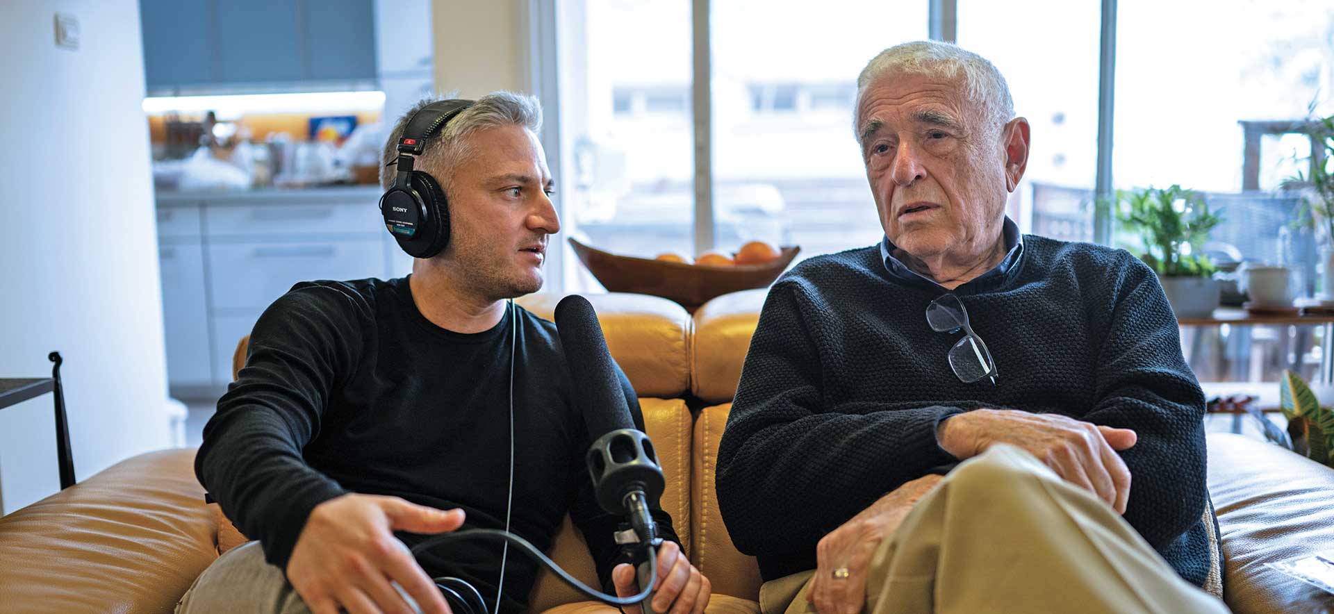 A person in headphones with microphone interviews another person. They are both sitting on a couch.