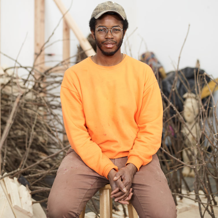 Artist Hugh Hayden wearing an orange sweater, a baseball hat, and glasses in his studio.